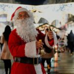 A joyful Santa Claus stands in a lively urban street in Belgrade at night, ringing a bell during Christmas time.
