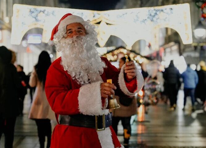 A joyful Santa Claus stands in a lively urban street in Belgrade at night, ringing a bell during Christmas time.