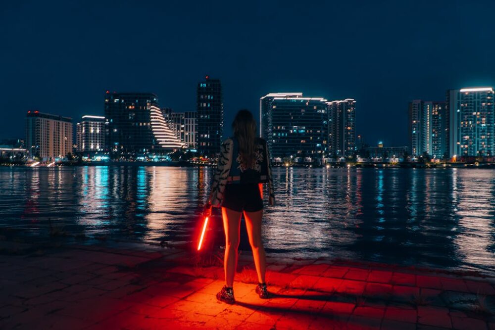 A woman with a red light stands by the water, overlooking Belgrade's illuminated skyline at night.