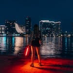 A woman with a red light stands by the water, overlooking Belgrade's illuminated skyline at night.