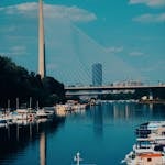 View of Ada Bridge in Belgrade with boats docked on the Sava River under a clear blue sky.